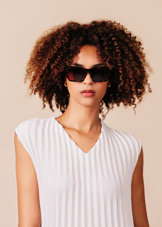 Woman wearing sunglasses and a white sleeveless top against a beige background