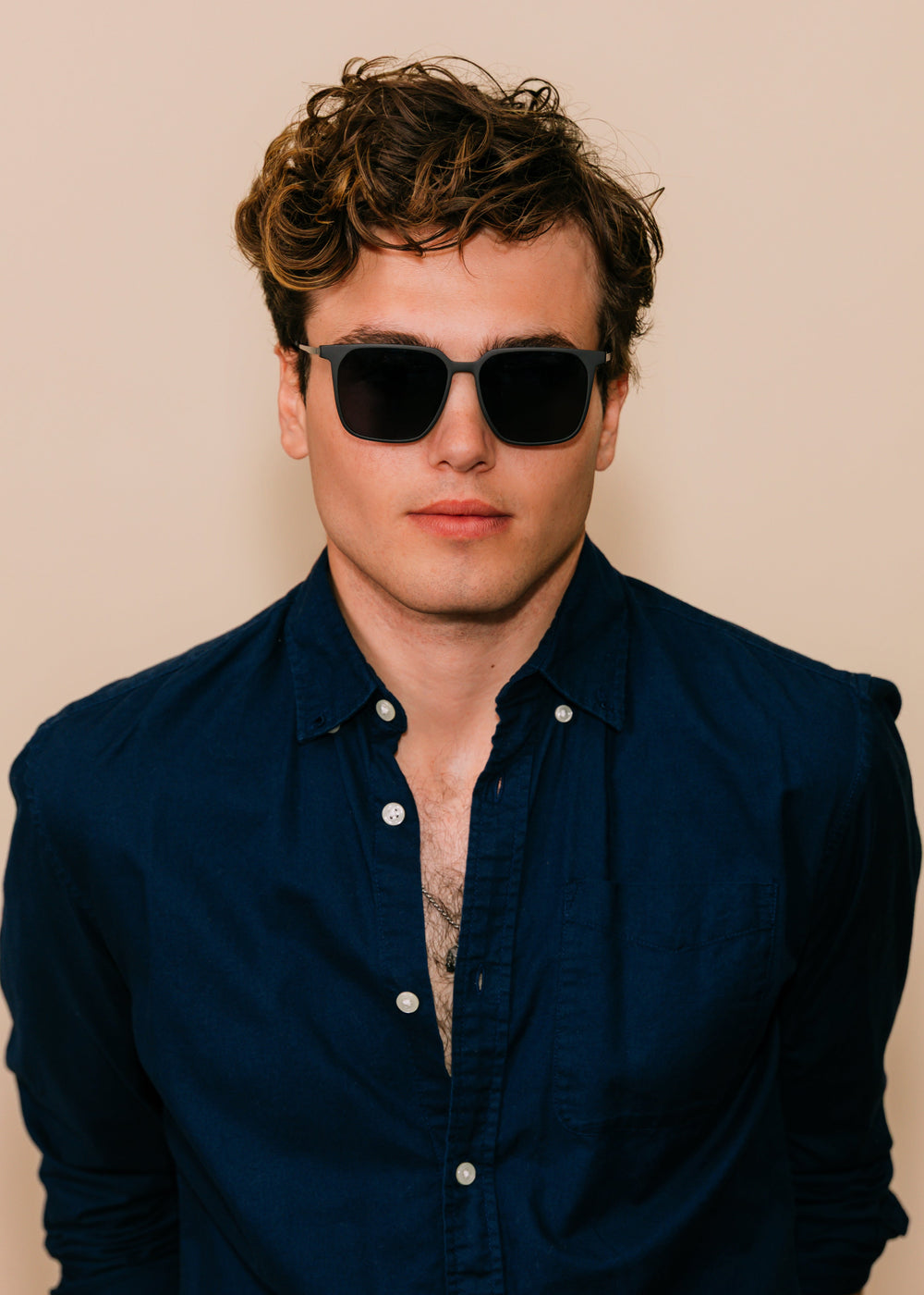 Man wearing sunglasses and a dark blue shirt against a beige background