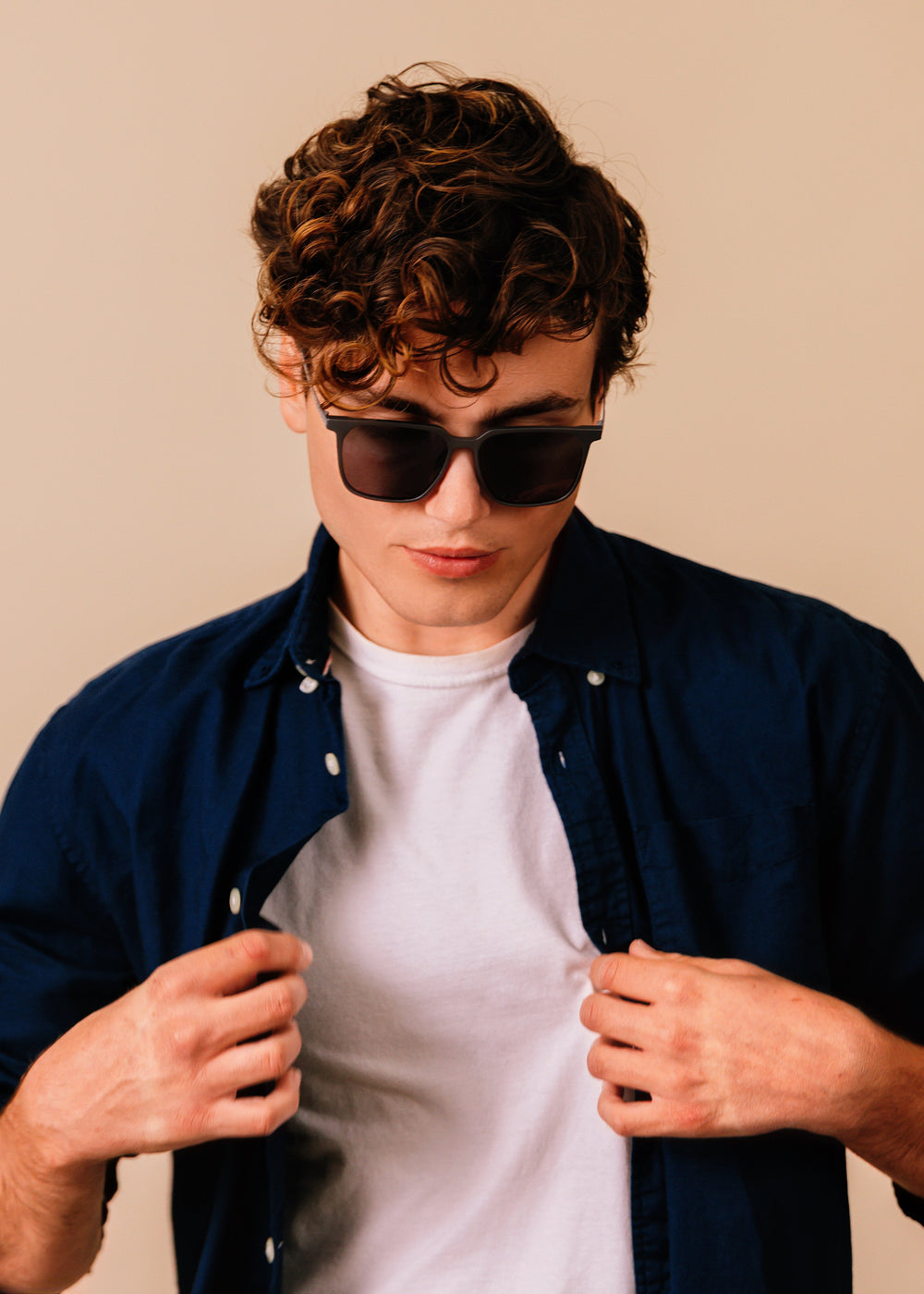 Man wearing sunglasses and a navy shirt over a white t-shirt against a beige background
