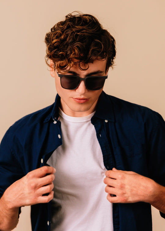 Man wearing sunglasses and a navy shirt over a white t-shirt against a beige background