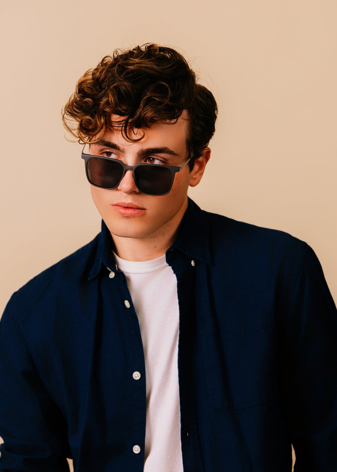 Man wearing sunglasses and a dark jacket over a white shirt on a beige background