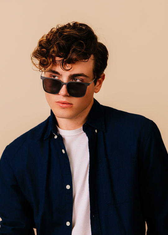 Man wearing sunglasses and a dark jacket over a white shirt on a beige background