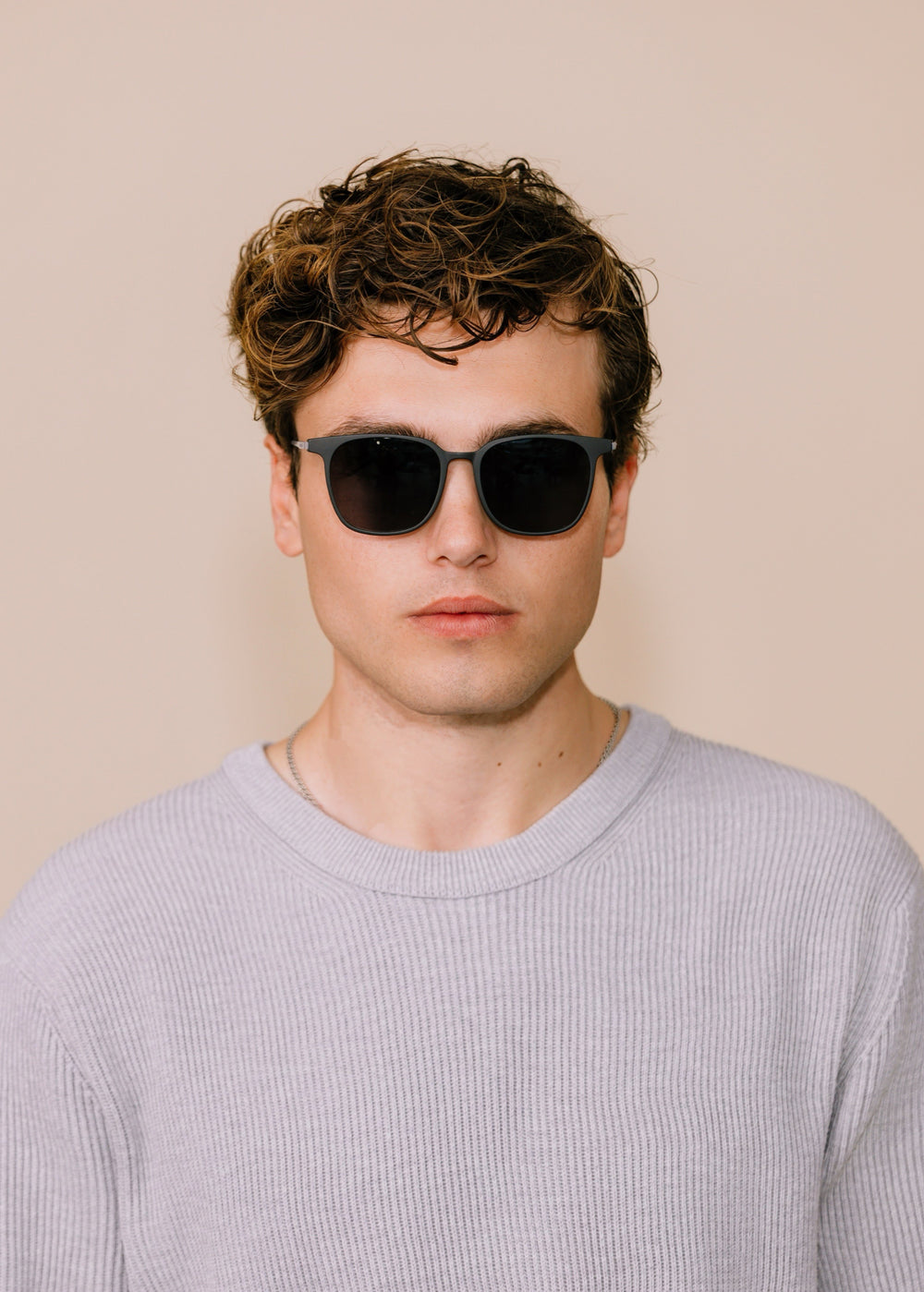 Man wearing sunglasses and a light gray sweater against a beige background