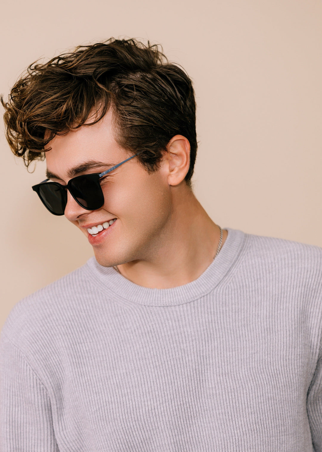 Male model wearing Achernar sunglasses in Classic Black with a light grey knit, turned to the side and smiling. 