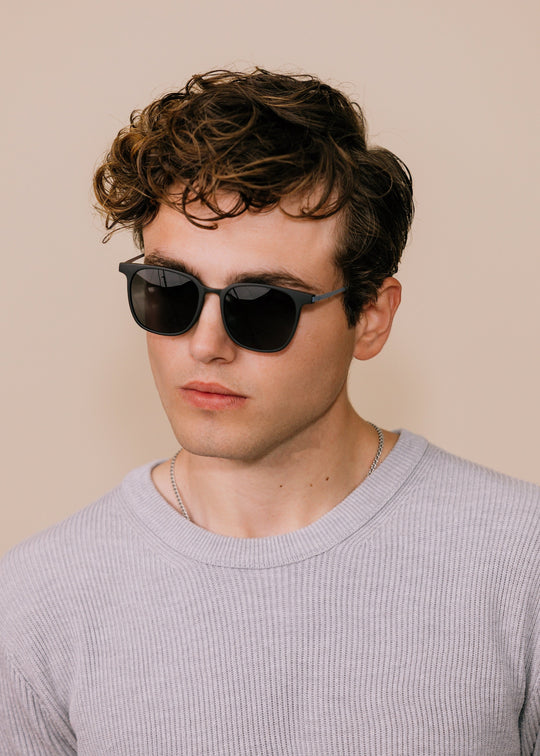 Male model wearing Achernar sunglasses in Classic Black with a light grey knit, head tilted down. 