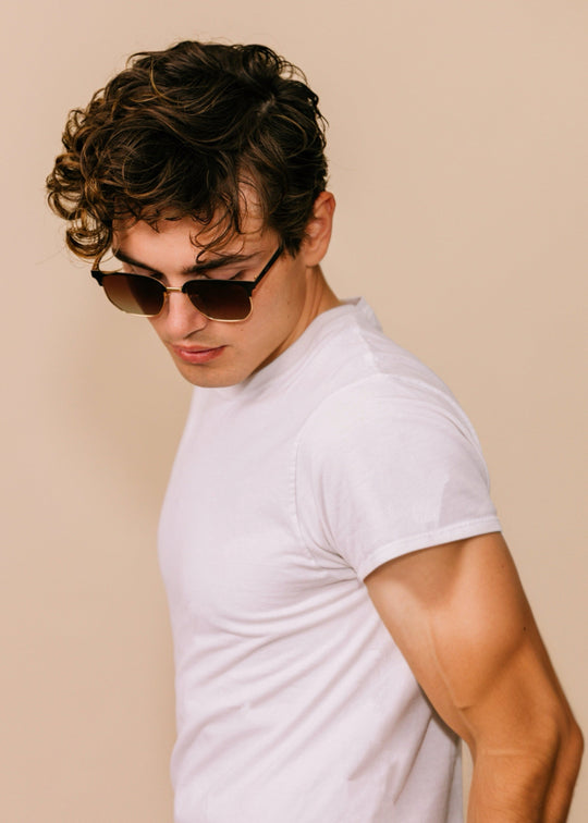 Three-quarter portrait of a model wearing Altessa sunglasses in Antique Brass with a white T-shirt against a neutral beige background. 