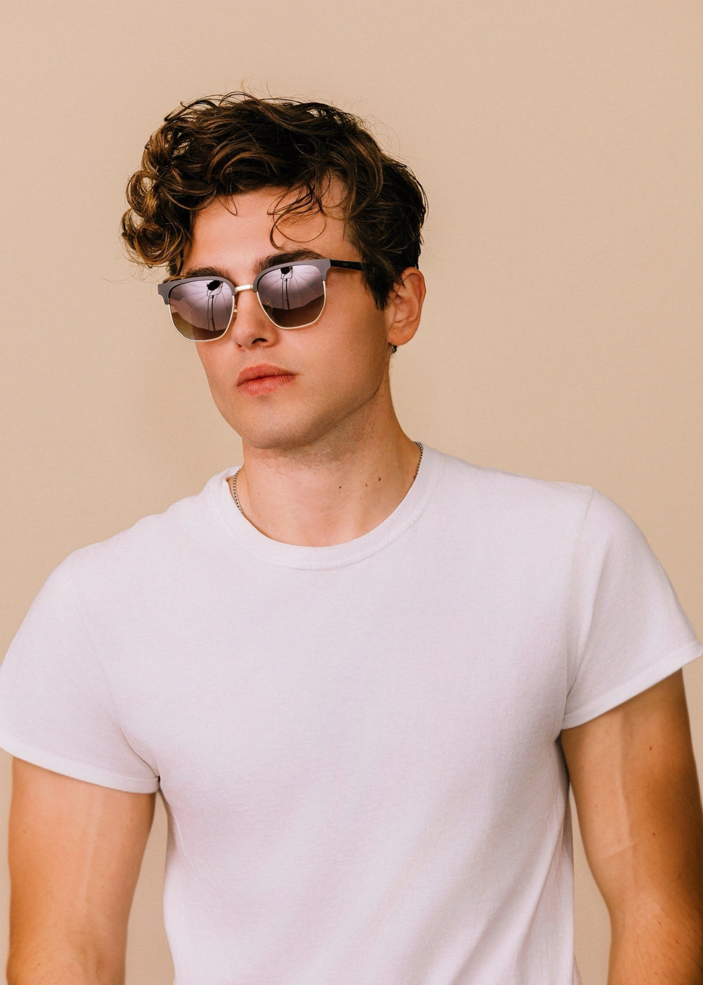 Front portrait of a model wearing Altessa sunglasses in Antique Brass with a white T-shirt against a neutral beige background.