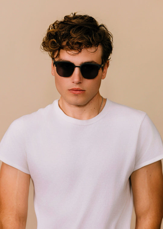 Front portrait of a model wearing Altessa sunglasses in Eclipse Black with a white T-shirt against a neutral beige background. 