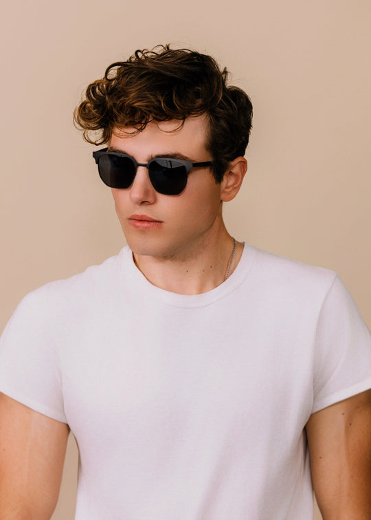 Three-quarter portrait of a model wearing Altessa sunglasses in Eclipse Black with a white T-shirt against a neutral beige background. 