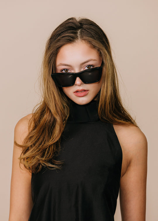 Female model wearing Aquila Black sunglasses with a black sleeveless high-neck top, head tilted with hair swept forward. 