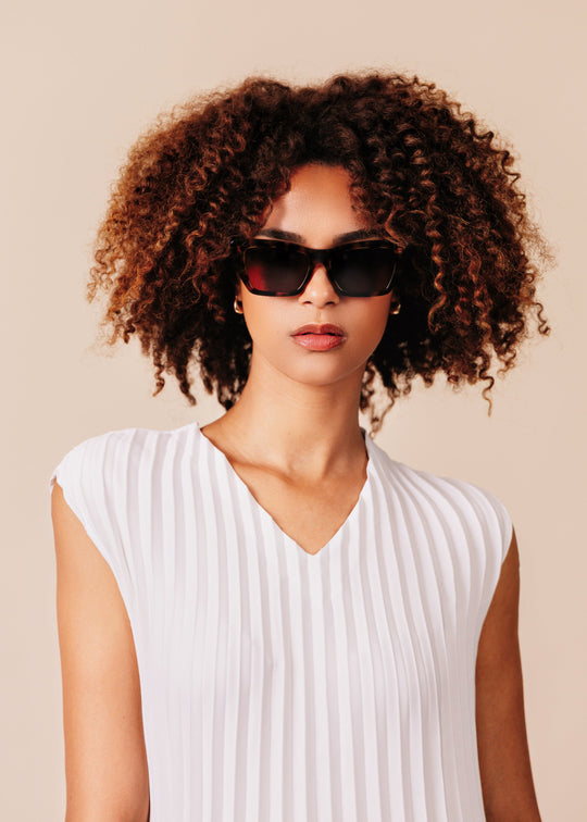 Female model wearing Aquila Tortoiseshell sunglasses with a white pleated sleeveless top, turned three-quarters with chin raised. 