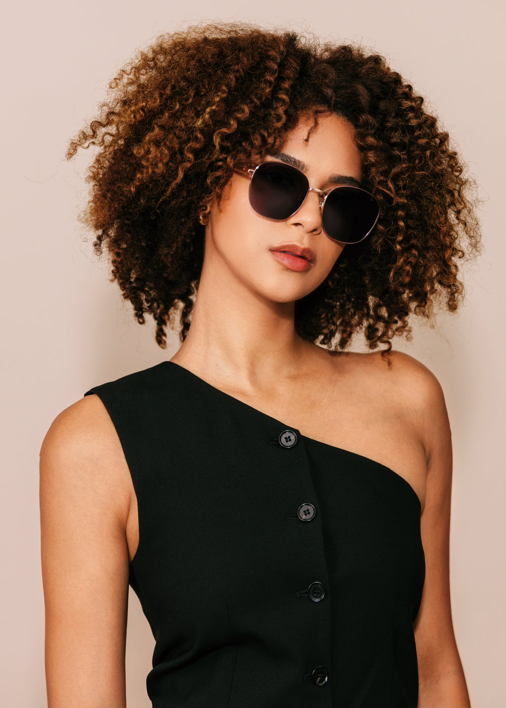 Woman wearing sunglasses and a black one-shoulder dress against a beige background