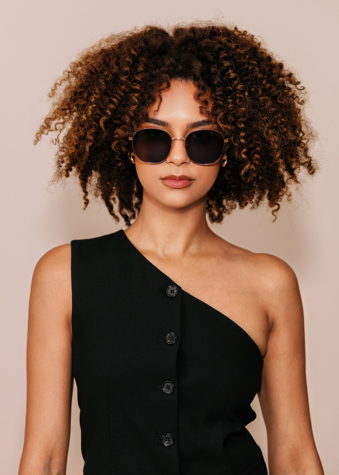 Woman wearing sunglasses and a black one-shoulder dress against a beige background