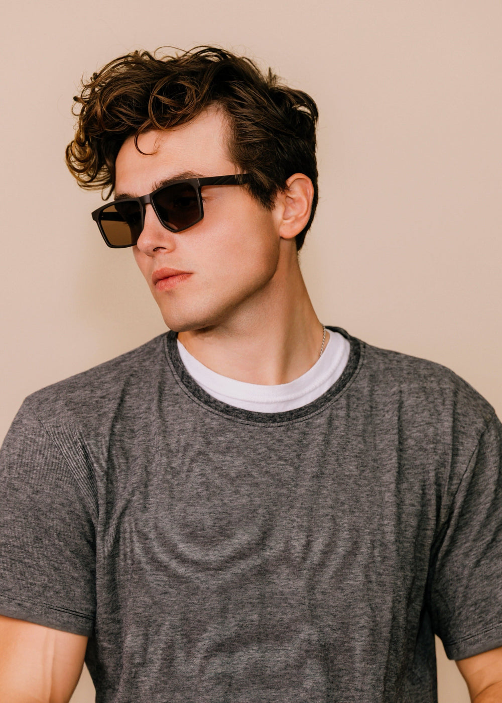 Male model wearing Betelgeuse matte brown sunglasses with a grey T-shirt, turned three-quarters to show the matte TR90 frame and warm brown lenses.
