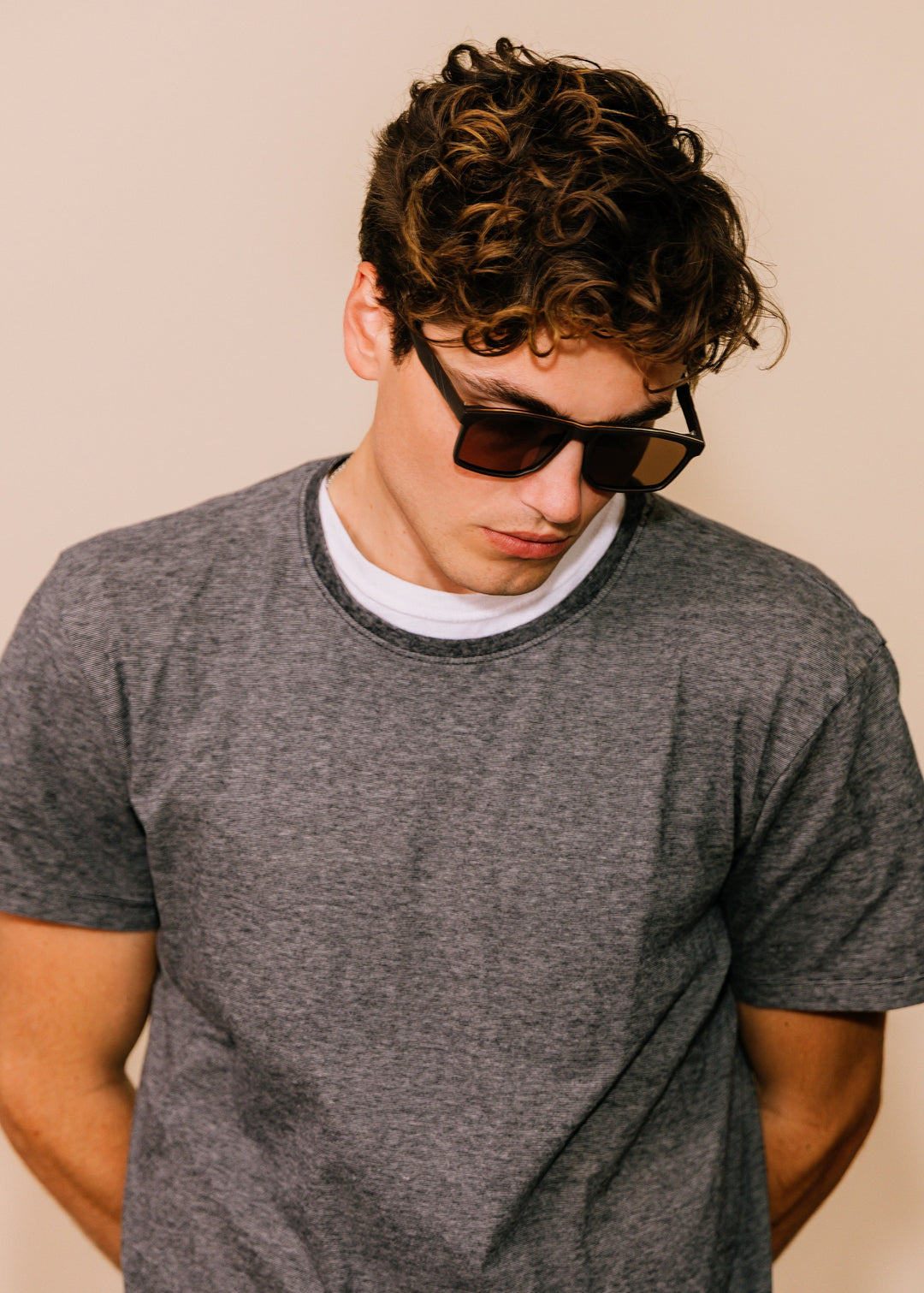 Male model wearing Betelgeuse matte brown sunglasses with a grey T-shirt, looking down to show how the frame sits on the face. 