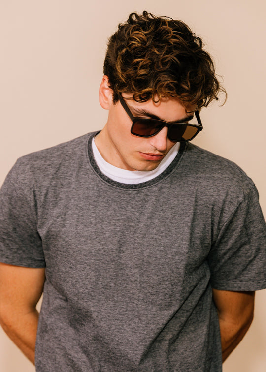 Male model wearing Betelgeuse matte brown sunglasses with a grey T-shirt, looking down to show how the frame sits on the face. 