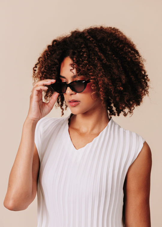 Female model wearing Chara sunglasses in Black with a white ribbed sleeveless top, one hand adjusting the temple. 