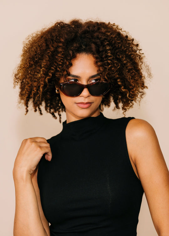 Three-quarter portrait of a model wearing Eclipse Feline sunglasses in Burnished Umber with a black sleeveless top against a neutral beige background. 