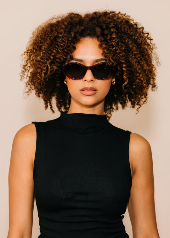 Front portrait of a model wearing Eclipse Feline sunglasses in Burnished Umber with a black sleeveless top against a neutral beige background. 