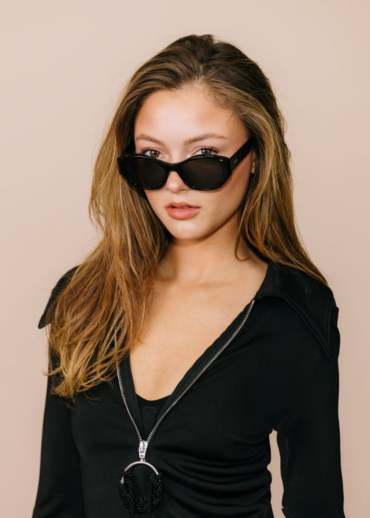 Three-quarter portrait of a model wearing Eclipse Feline sunglasses in Obsidian Black with a black outfit against a neutral beige background. 
