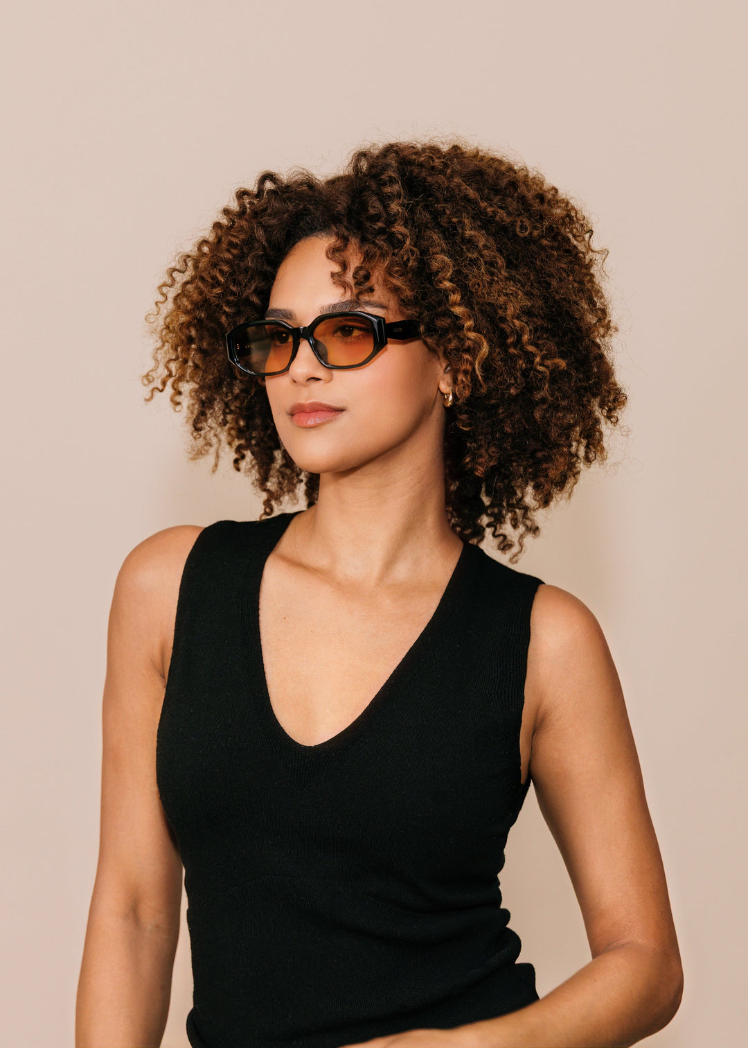 Female model wearing Lyra sunglasses in Amber with a black sleeveless top, turned slightly to one side. 