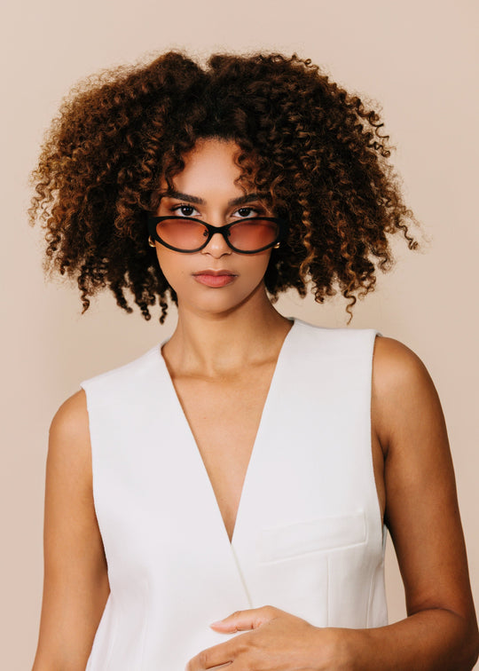 Front portrait of a model wearing Nyx sunglasses in Blush Eclipse with a white sleeveless top against a neutral beige background. 