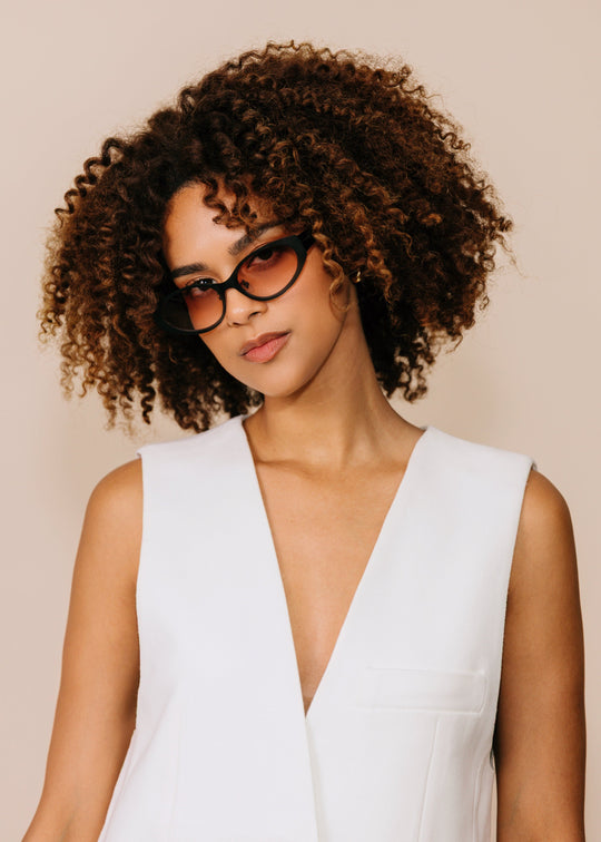 Three-quarter portrait of a model wearing Nyx sunglasses in Blush Eclipse with a white sleeveless top against a neutral beige background. 