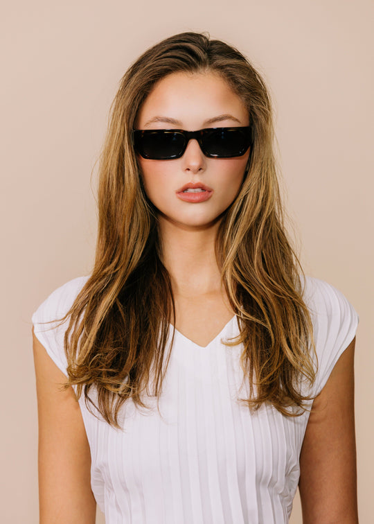 Woman wearing black sunglasses and a white top against a beige background 