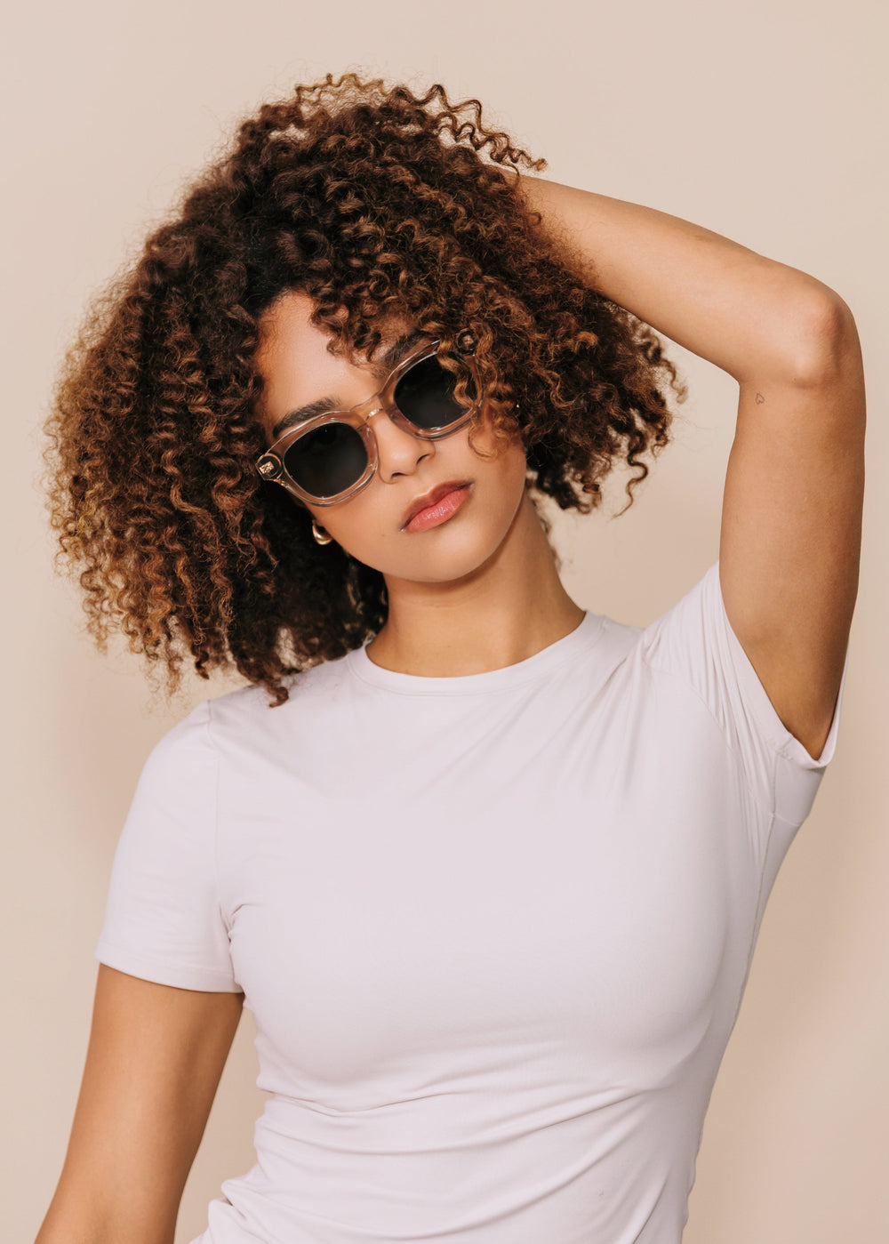 Model wearing Phantom Verge sunglasses in Champagne Moss, turned three-quarter with one hand raised to her hair, showing the translucent champagne frame and deep green lenses.