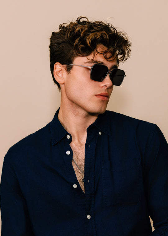 Male model wearing Rigel sunglasses in Gunmetal Ash with a navy button-down shirt, turned three-quarters. 