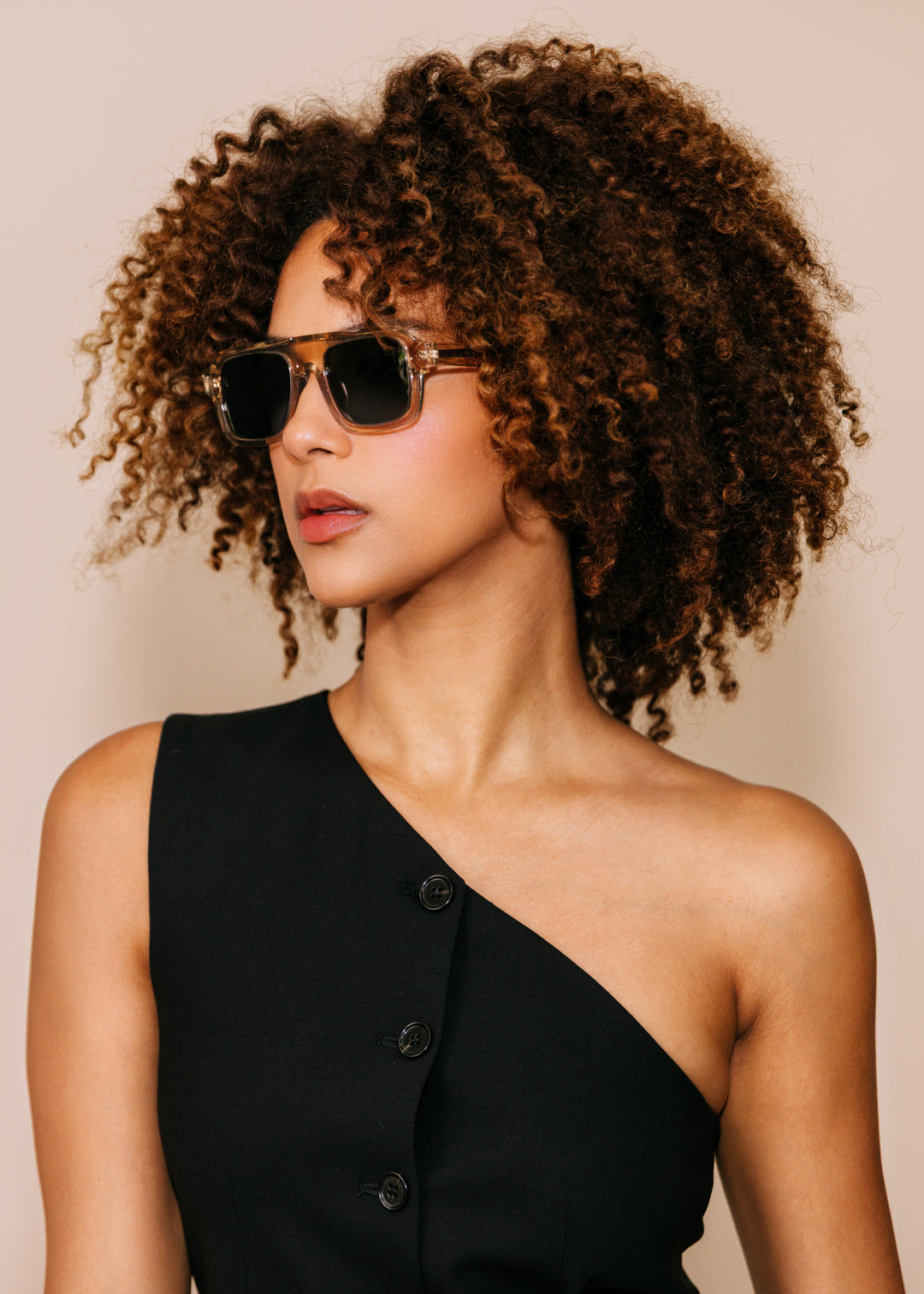 Woman wearing sunglasses and a black one-shoulder dress against a beige background