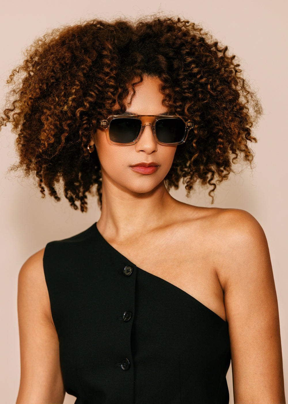 Woman wearing sunglasses and a black one-shoulder dress against a beige background