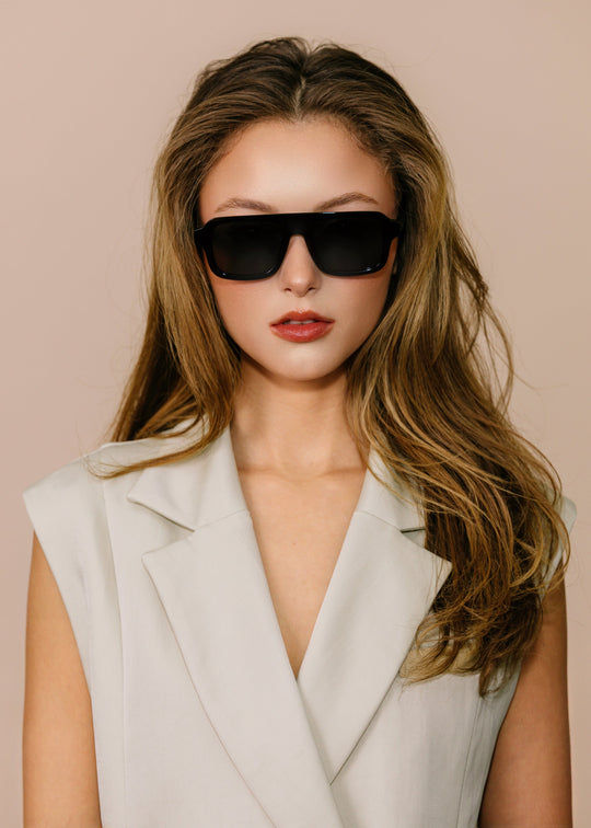 Woman wearing sunglasses and a light-colored sleeveless blazer against a beige background 