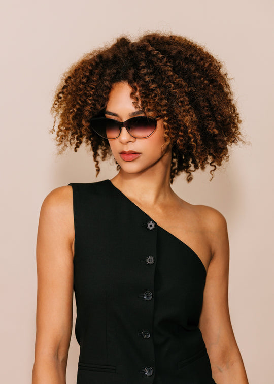 Woman wearing a black one-shoulder dress and sunglasses against a beige background