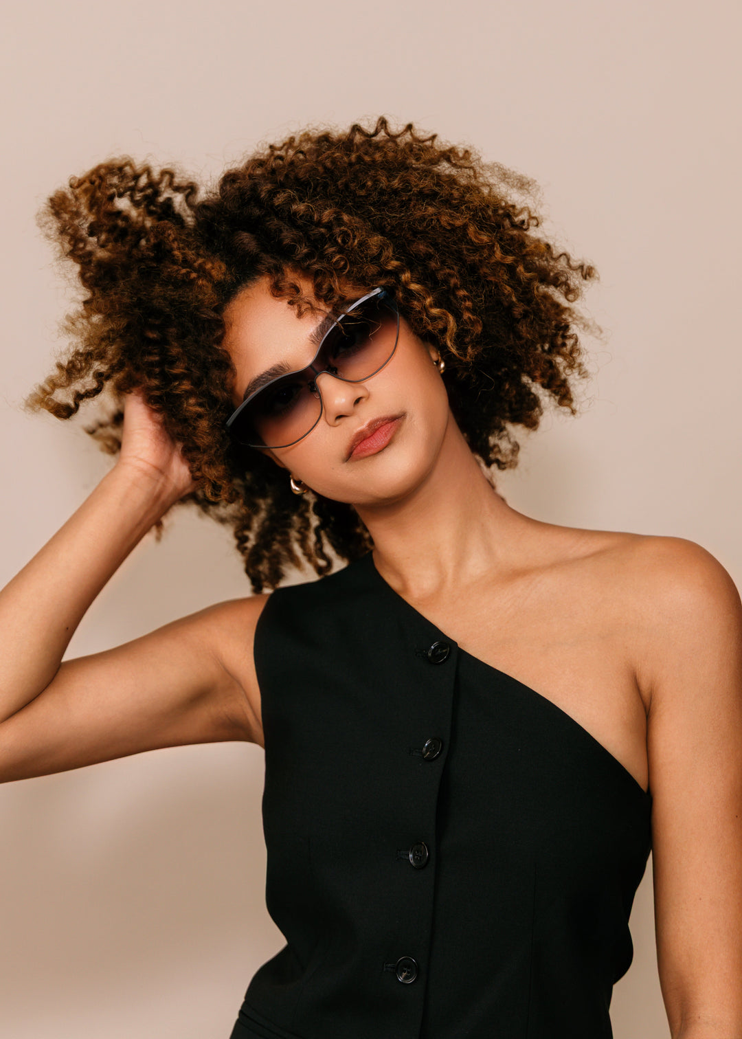 Woman wearing a black one-shoulder dress and sunglasses against a beige background
