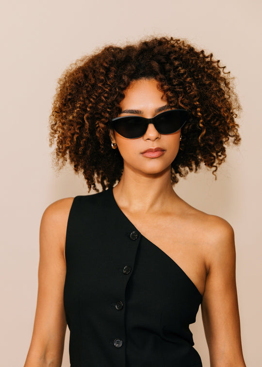 Model wearing Zenith sunglasses in Onyx Black with a black one-shoulder dress against a neutral beige background. 