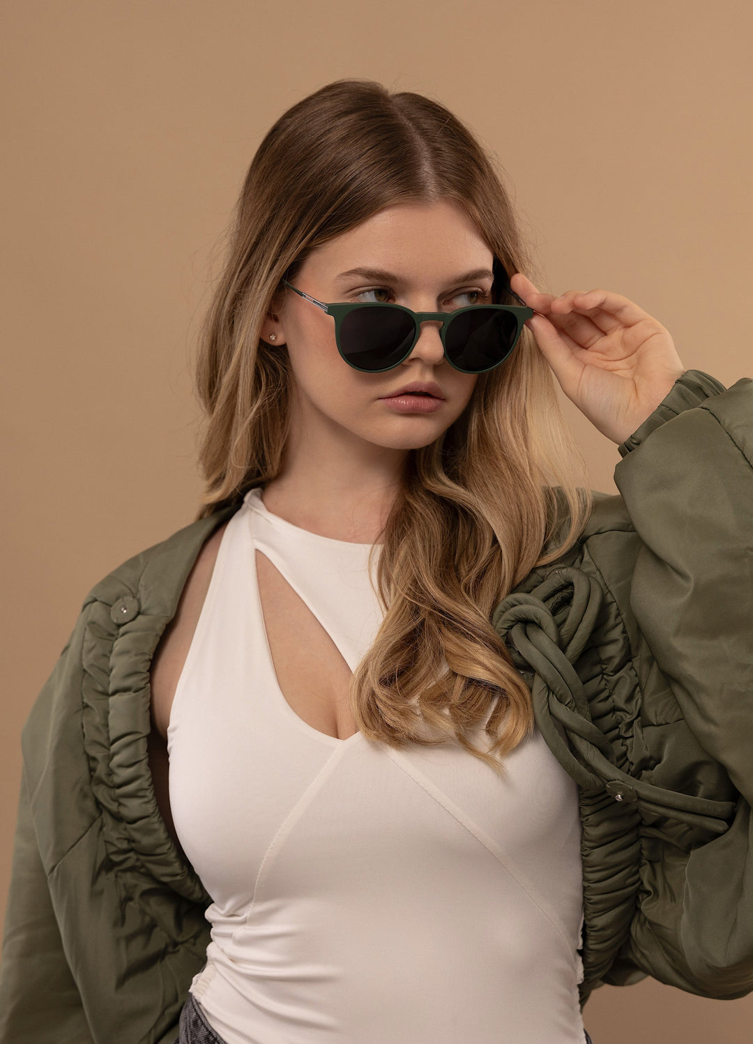 Female model wearing Altair sunglasses in Matte Green with an olive bomber jacket over a white top, one hand raised to her hair. 