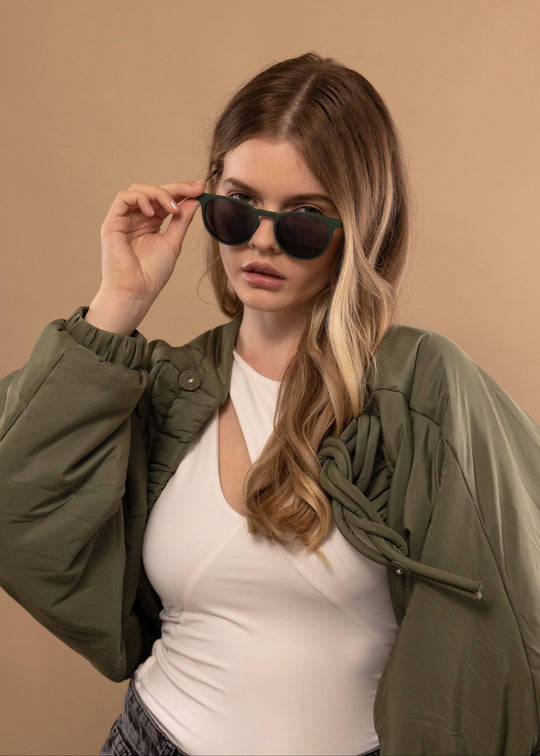 Female model wearing Altair sunglasses in Matte Green with an olive bomber jacket over a white top, one hand adjusting the temple. 
