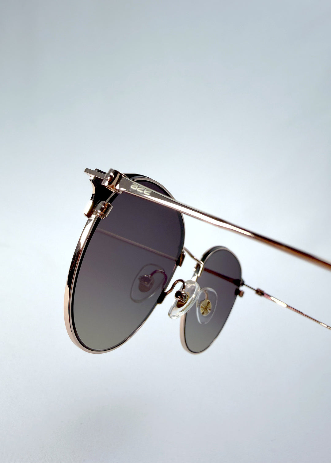 Close-up hinge detail view of AZE Aster Line sunglasses in Duskgold, featuring a gold-tone rounded frame and plum-to-amber gradient lenses, showing the hinge construction, lens edge, and temple connection.