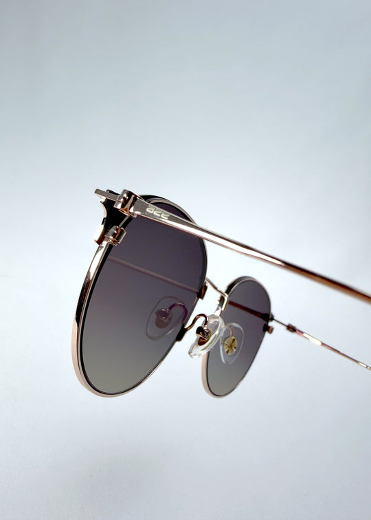 Close-up hinge detail view of AZE Aster Line sunglasses in Duskgold, featuring a gold-tone rounded frame and plum-to-amber gradient lenses, showing the hinge construction, lens edge, and temple connection.