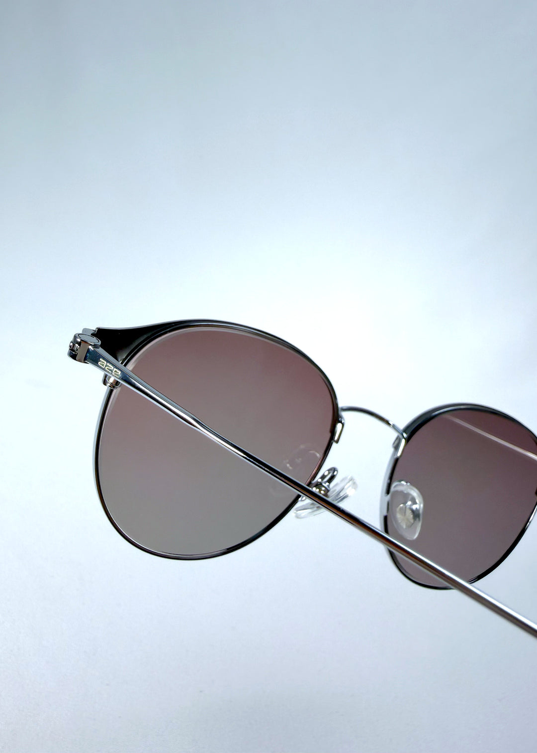 Close-up hinge detail view of AZE Aster Line sunglasses in Mocha Dusk, featuring a silver-tone rounded frame and taupe gradient lenses, showing the hinge construction, lens edge, and temple connection.