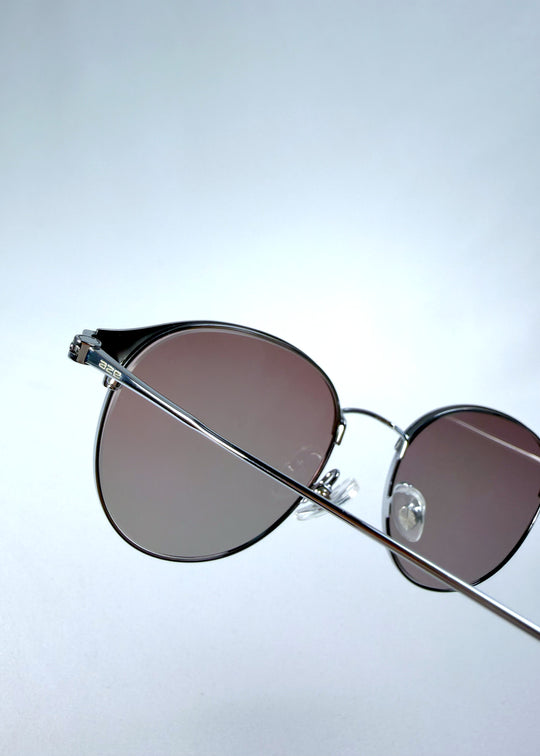 Close-up hinge detail view of AZE Aster Line sunglasses in Mocha Dusk, featuring a silver-tone rounded frame and taupe gradient lenses, showing the hinge construction, lens edge, and temple connection.