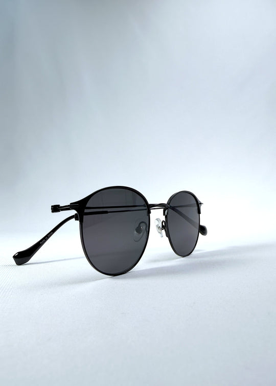Folded view of AZE Aster Line sunglasses in Obsidian Black, featuring a dark geometric round frame and smoky gray lenses, showing the folded temples and frame profile.
