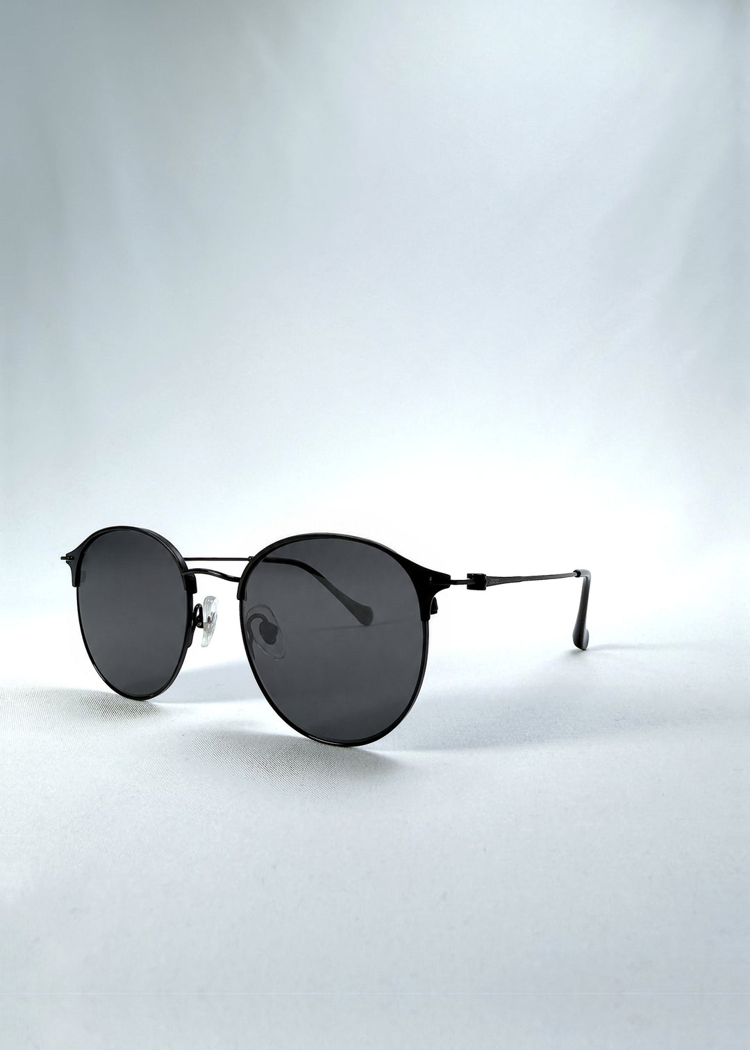 Three-quarter front view of AZE Aster Line sunglasses in Obsidian Black, featuring a dark geometric round frame and smoky gray lenses, showing the frame shape and temple profile.