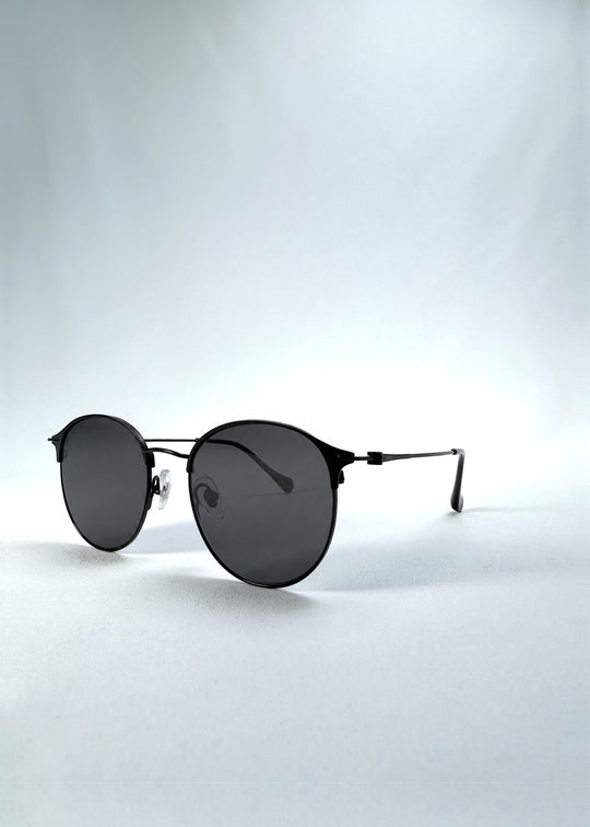 Three-quarter front view of AZE Aster Line sunglasses in Obsidian Black, featuring a dark geometric round frame and smoky gray lenses, showing the frame shape and temple profile.