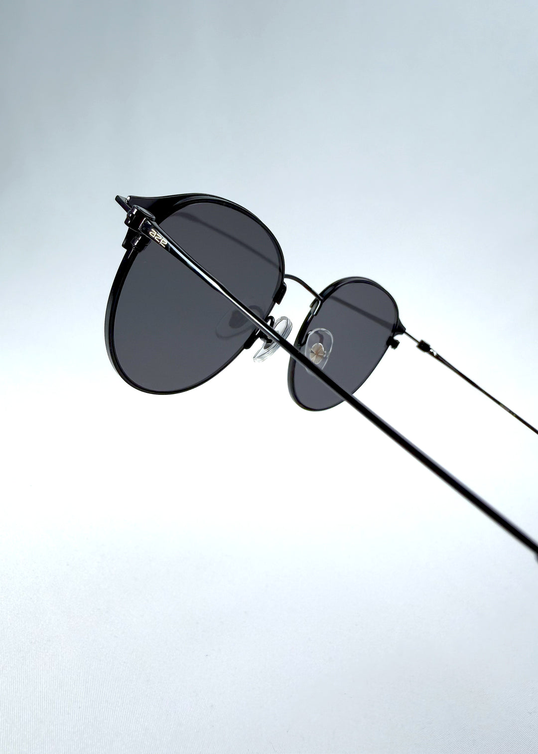 Close-up hinge detail view of AZE Aster Line sunglasses in Obsidian Black, featuring a dark geometric round frame and smoky gray lenses, showing the hinge construction, lens edge, and temple connection.