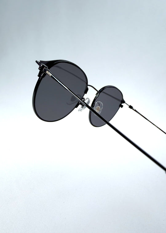 Close-up hinge detail view of AZE Aster Line sunglasses in Obsidian Black, featuring a dark geometric round frame and smoky gray lenses, showing the hinge construction, lens edge, and temple connection.