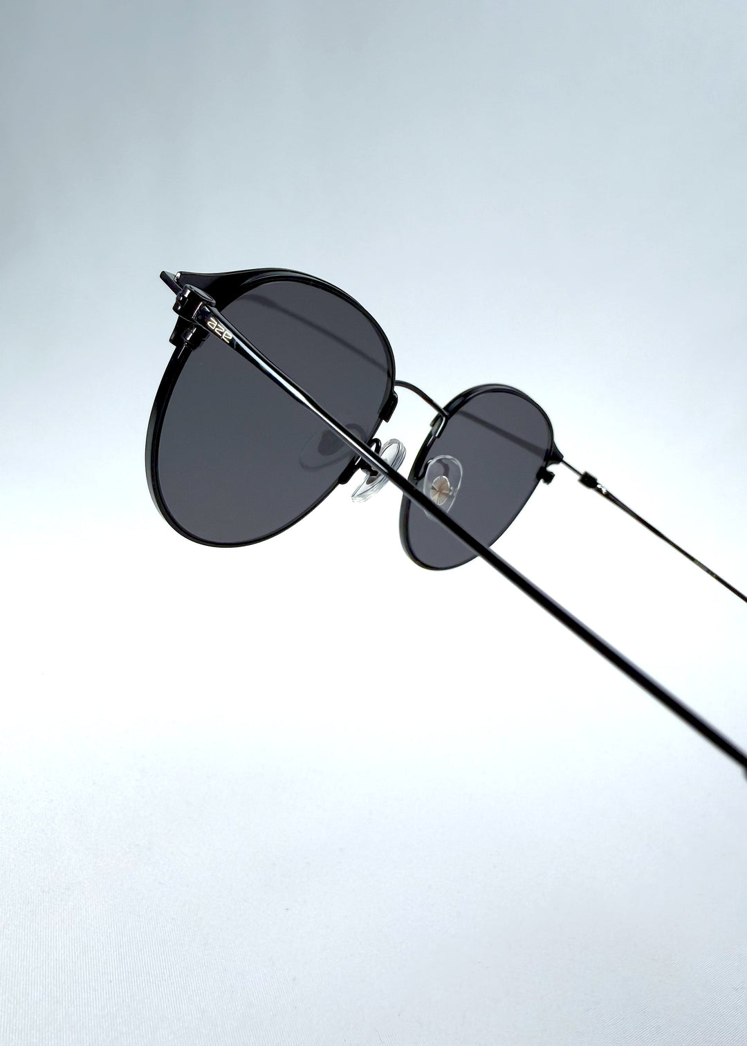 Close-up hinge detail view of AZE Aster Line sunglasses in Obsidian Black, featuring a dark geometric round frame and smoky gray lenses, showing the hinge construction, lens edge, and temple connection. 