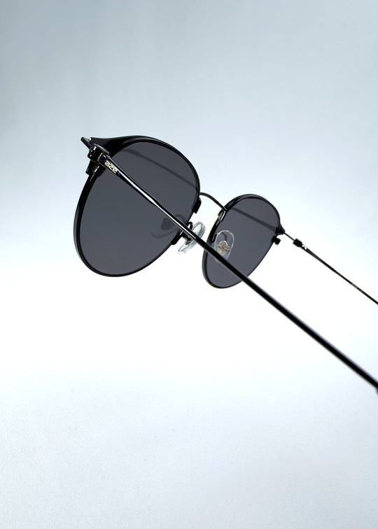 Close-up hinge detail view of AZE Aster Line sunglasses in Obsidian Black, featuring a dark geometric round frame and smoky gray lenses, showing the hinge construction, lens edge, and temple connection. 