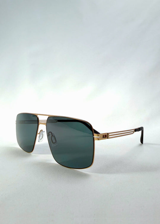 Three-quarter front view of Axiom sunglasses in Antique Brass, featuring antique brass rectangular metal frame and green-grey polarized lenses, showing the front shape and temple profile. 